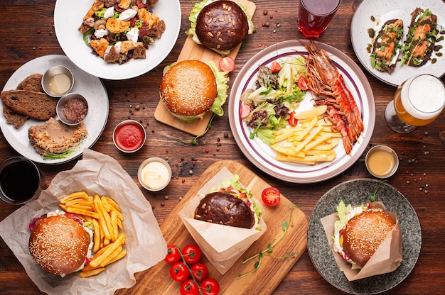 Burgers, french fries, salads, grilled shrimps, sauces, beer and other drinks served on the wooden table. Horizontal shot of top view.