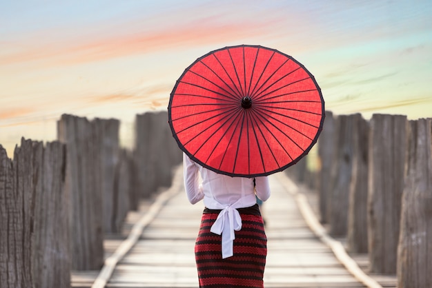 Burmese woman holding traditional red umbrella and walking on U Bein Bridge, Mandalay, Myanmar