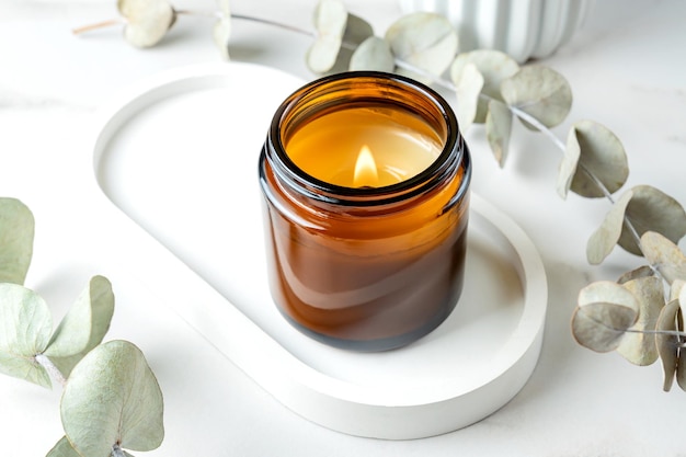 Burning candle in amber glass jar on concrete trendy tray and eucalyptus branches on white marble background close up