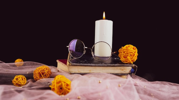 Burning candle books and thin veil with decor Composition of burning wax candle near eyeglasses on stack of books near thin fabric with decorative wicker balls on black background