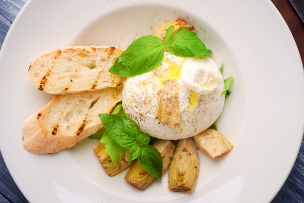 Burrata cheese with artichokes and Basil and a white plate on wood table, top view