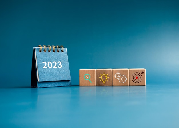 Business action plan with goal management idea and research icon signs on wooden cube blocks with small blue desk calendar year 2023 on blue background Business strategy concept minimal style