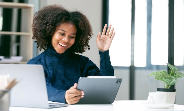 Business african woman having a video call on laptop waving hand and smiling to tablet screen sitting at office