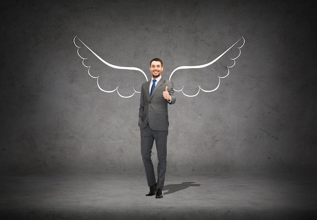 business, angel investor, safety, security and people concept - smiling young businessman with wings drawing showing thumbs up over gray concrete background