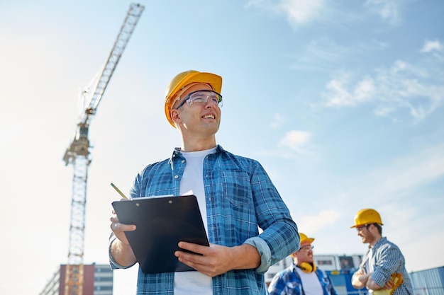 business, building, paperwork and people concept - happy builder in hardhat with clipboard and pencil over group of builders at construction site