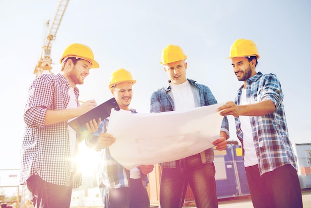 business, building, teamwork and people concept - group of smiling builders in hardhats with clipboard and blueprint outdoors