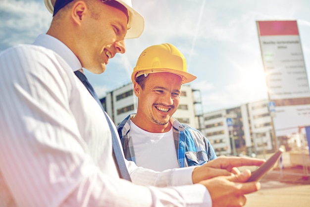 business, building, teamwork, technology and people concept - smiling builders in hardhats with tablet pc computer at construction