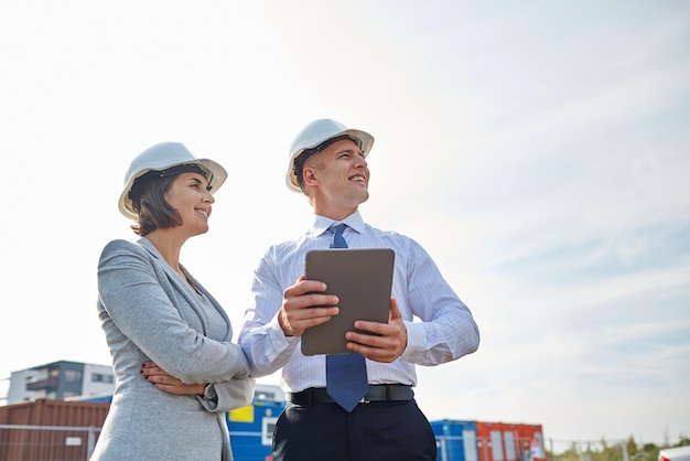 business, building, teamwork, technology and people concept - smiling man and woman in hardhats with tablet pc computer at construction site