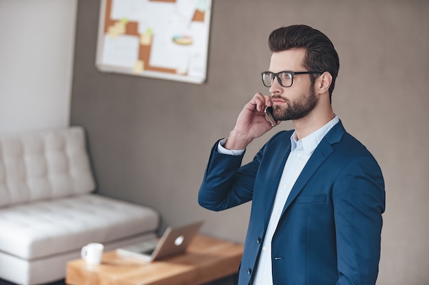 Business call. Handsome young man wearing glasses talking on mobile phone and looking away while standing in office