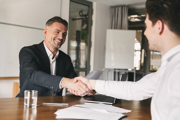 Business, career and placement concept - joyful handsome businessman 30s smiling and shaking hands with male candidate, who was recruited during interview in office