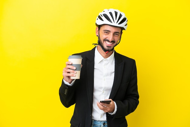 Business caucasian man with a bike helmet isolated on yellow background holding coffee to take away and a mobile