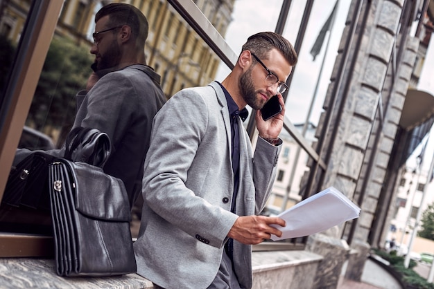 Business communications young businessman standing leaning on wall on the city street talking on