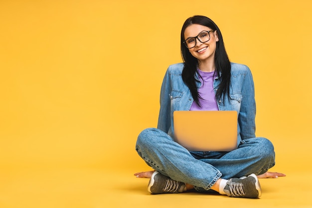 Business concept Portrait of happy young woman in casual sitting on floor in lotus pose and holding laptop isolated over yellow background