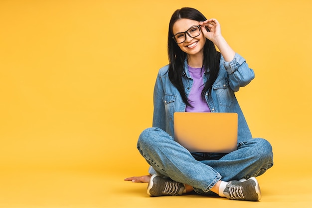 Business concept Portrait of happy young woman in casual sitting on floor in lotus pose and holding laptop isolated over yellow background