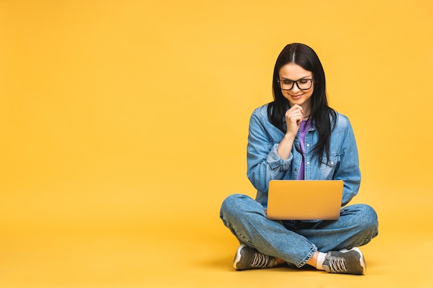 Business concept Portrait of happy young woman in casual sitting on floor in lotus pose and holding laptop isolated over yellow background