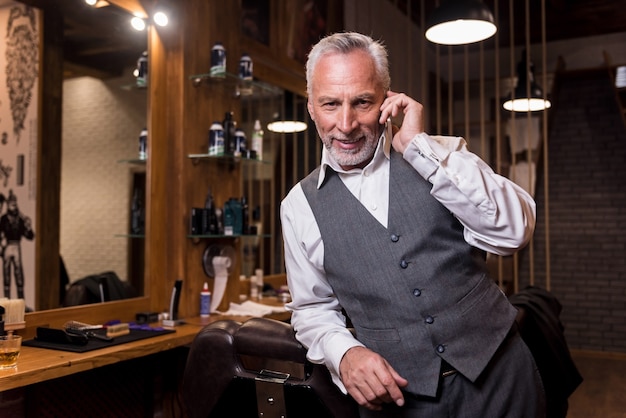 Business conversation. Handsome bearded senior man leaning on armchair and smiling while talking on mobile phone at barbershop.