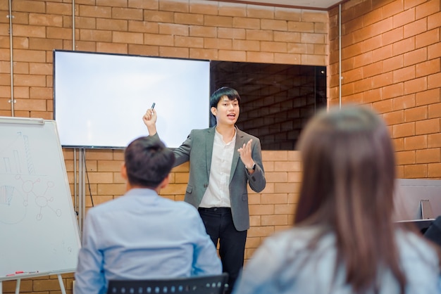 Business cooperation : Young asian male coach or speaker make flip chart presentation to diverse businesspeople at meeting in office. Male tutor or trainer present project to diverse colleagues.