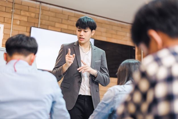 Business cooperation : Young asian male coach or speaker make flip chart presentation to diverse businesspeople at meeting in office. Male tutor or trainer present project to diverse colleagues.