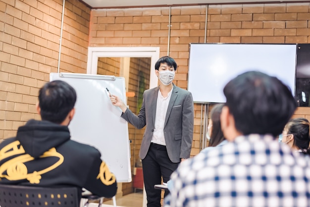 Business cooperation : Young asian male coach or speaker make flip chart presentation to diverse businesspeople at meeting in office. Male tutor or trainer present project to diverse colleagues.