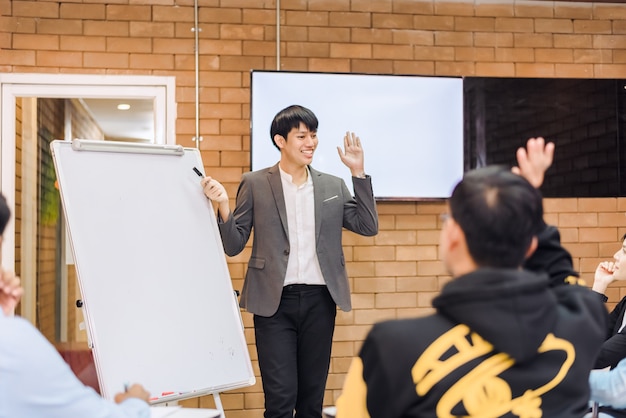 Business cooperation : Young asian male coach or speaker make flip chart presentation to diverse businesspeople at meeting in office. Male tutor or trainer present project to diverse colleagues.