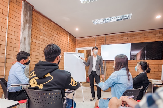 Business cooperation : Young asian male coach or speaker make flip chart presentation to diverse businesspeople at meeting in office. Male tutor or trainer present project to diverse colleagues.