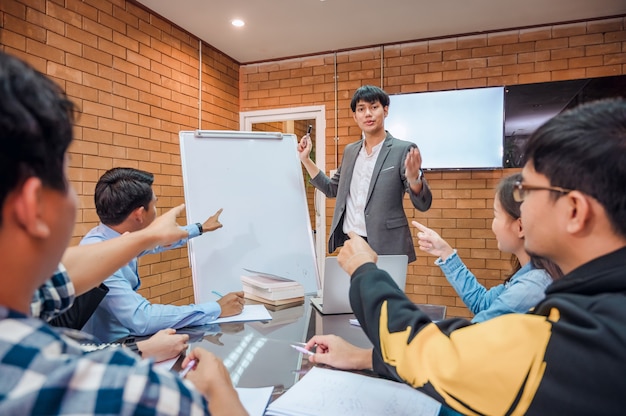 Business cooperation : Young asian male coach or speaker make flip chart presentation to diverse businesspeople at meeting in office. Male tutor or trainer present project to diverse colleagues.