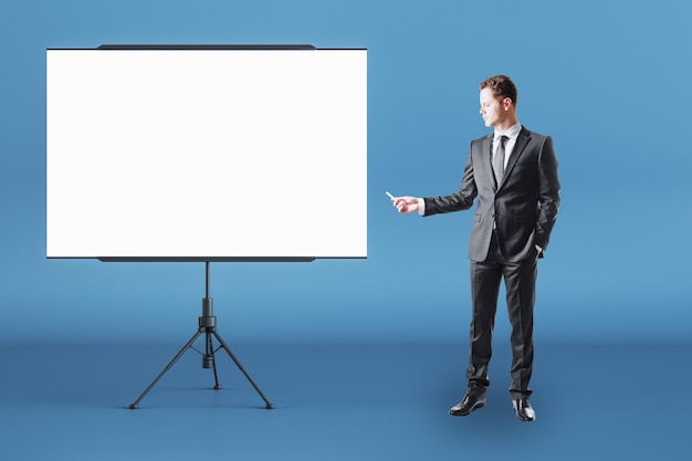 Business education concept with businessman front view with chalk in right hand near blank white flip chart with place for your logo or text in abstract room on blue wall background mock up