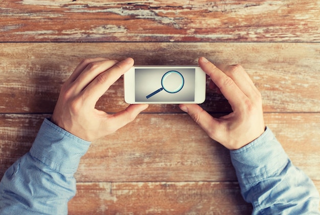 business, education, people and technology concept - close up of male hands holding smartphone with magnifying glass picture on screen at table