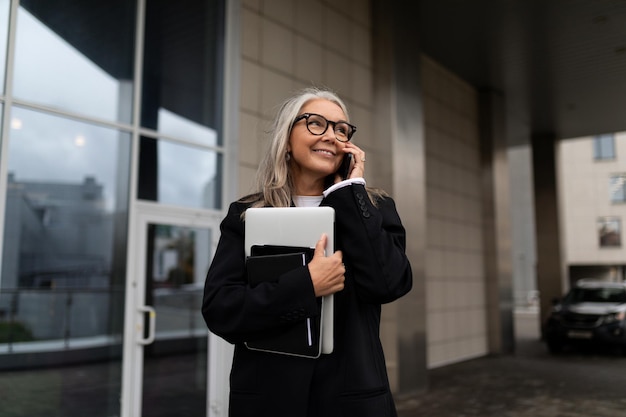 A business elderly woman speaks on a mobile phone with a laptop in her hands against the backdrop of