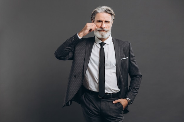 Business and fashionable bearded mature man in a gray suit holding his mustache on the grey wall.