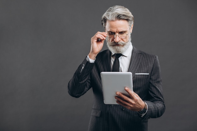 Business and fashionable bearded mature man in a gray suit making shopping in tablet with his gold credit card on the grey wall.