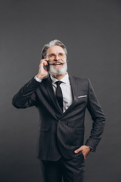 Business and fashionable bearded mature man in a gray suit smiling and talking by phone on the grey wall.