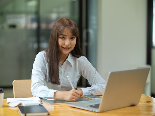 business female smiling and working on tablet while hold stylus pen with laptop at office