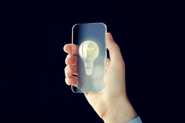 business, future technology, idea, startup and people concept - close up of male hand holding and showing transparent smartphone with lightbulb icon on screen over black background