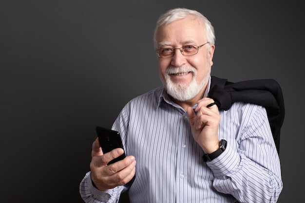 Business-haired, white-haired man in glasses with a phone on a gray background smiles friendly