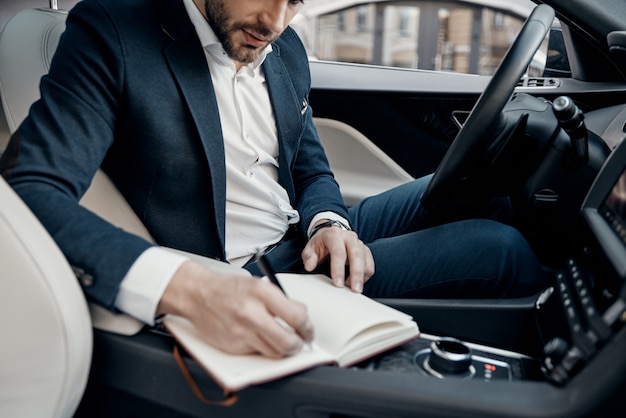 Business is his life. Close up of young man in full suit writing something down in personal organizer while sitting inside of the car