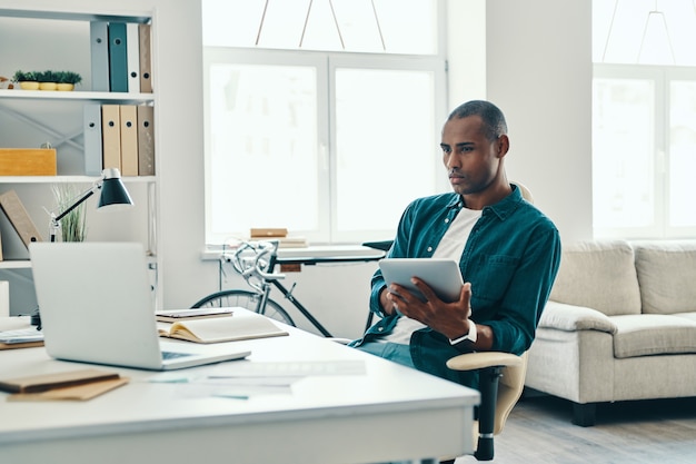 Business is his life. Thoughtful young African man in shirt working using digital tablet while sitting in the office