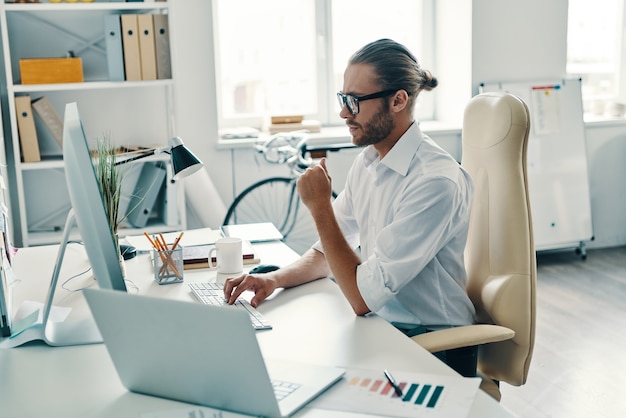 Business is his life. Thoughtful young man in shirt working using computer while sitting in the office