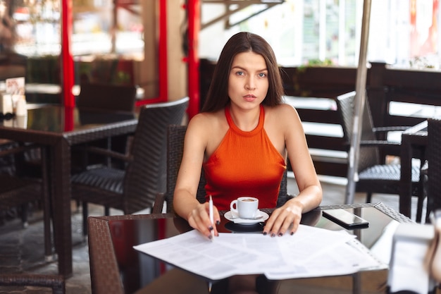 Business lady girl sits at a table in a cafe, considers paper, thinks. Resume, signing an important business deal. Work away from home.