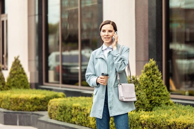 Business lady in trench coat talking on phone and drinks coffee on the go in city Lifestyle