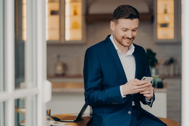 Business lifestyle. Glad handsome man with stubble uses mobile phone for texting online checks newsfeed wears elegant clothes posesagainst blurred coworking space. Focused salesman with gadget