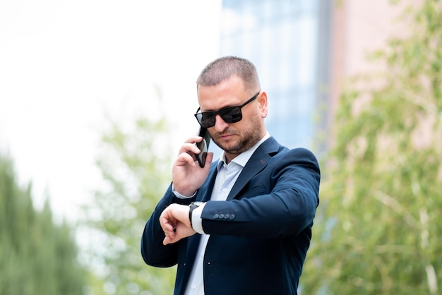 A business man in dark sunglasses stands outside the business building talks on the phone.