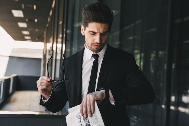 Business man near business center looking holding newspaper looking at watch.