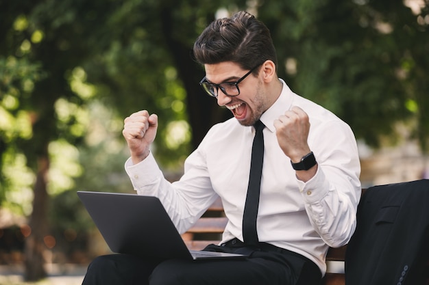 Business man outdoors in the park using laptop computer make winner gesture.