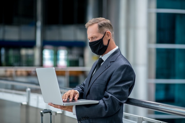 Business man. Serious adult man in business suit and protective mask working at laptop waiting for flight at airport