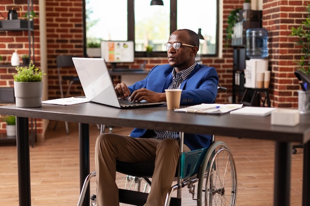 Business man sitting in wheelchair working on laptop to plan project and strategy in office. Person with disability using computer to work on company startup and do management research.