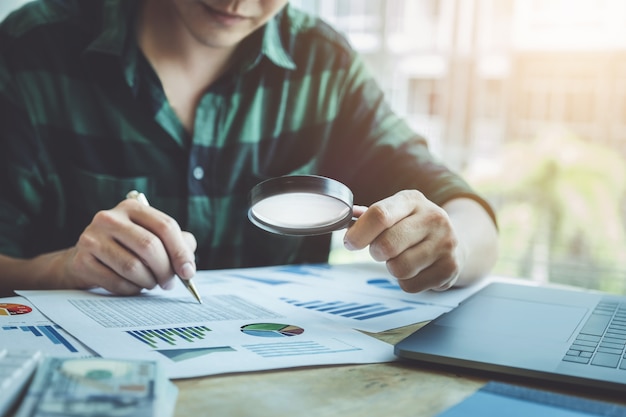 Business man using magnifying to review balance sheet annual with using laptop computer to calculating budget. audit and Check integrity before investment concept.