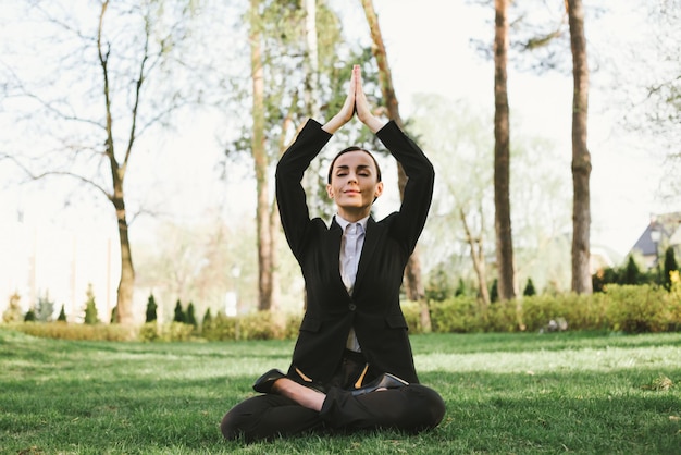 Business and meditation A concentrated beautiful business woman in a black suit is sitting in a lotus position on the grass in the city park and resting after work