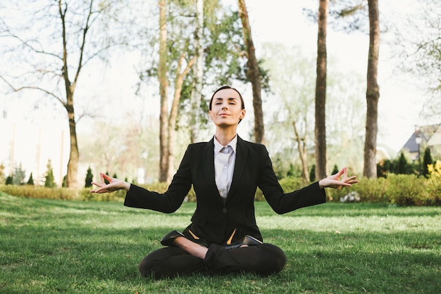 Business and meditation A concentrated beautiful business woman in a black suit is sitting in a lotus position on the grass in the city park and resting after work