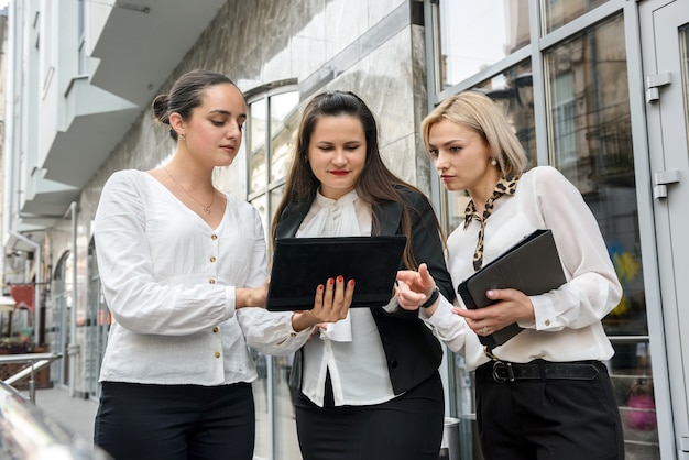 Business meeting outside building. Three young ladies in business suits discussing information on tablet